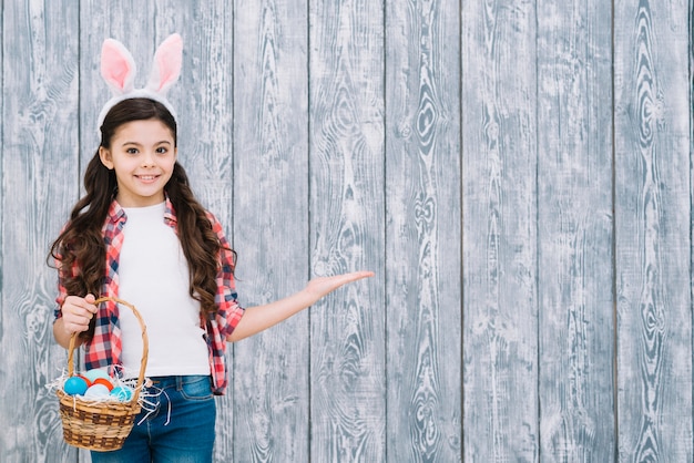 Menina sorridente, com, orelhas bunny, segurando, cesta, ovos páscoa, apresentando, contra, cinzento, escrivaninha madeira