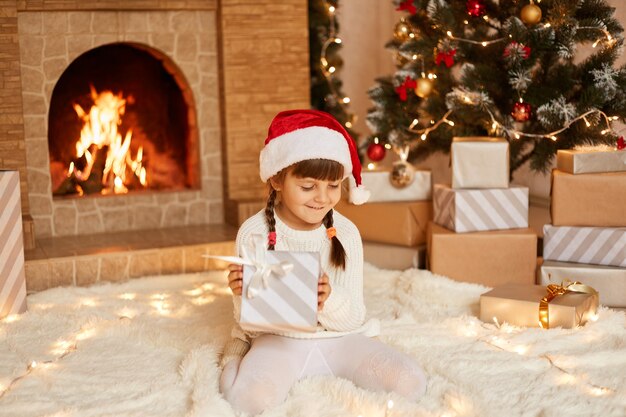 Menina sorridente com suéter branco e chapéu de Papai Noel, sentada no chão perto da árvore de Natal, caixas de presentes e lareira, segurando um presente dos pais nas mãos.
