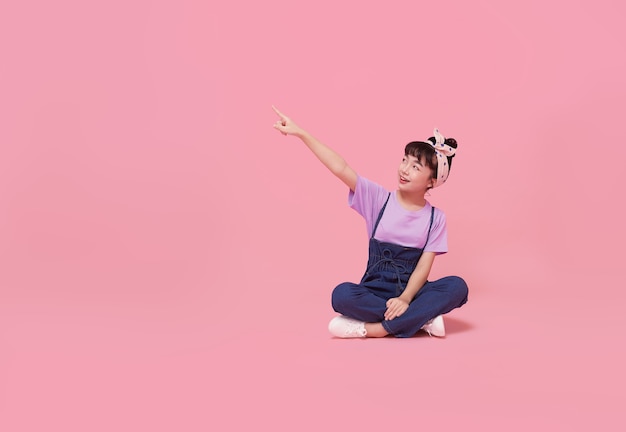 Menina sorridente criança asiática apontando o dedo no espaço em branco ao lado na parede rosa isolada.