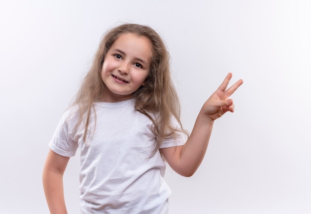 Menina sorridente da escola usando uma camiseta branca mostrando um gesto de paz em um fundo branco isolado
