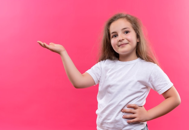 Foto grátis menina sorridente da escola vestindo uma camiseta branca aponta para o lado da mão no quadril em fundo rosa isolado