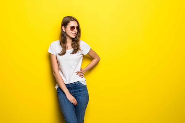 Menina sorridente da moda em camiseta branca e calça jeans azul fica na frente do fundo amarelo do estúdio