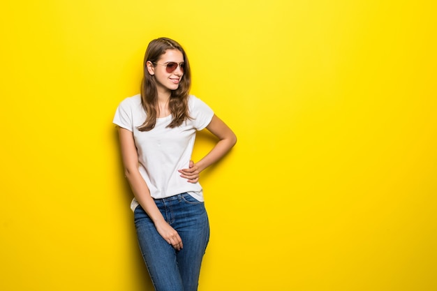 Menina sorridente da moda em camiseta branca e calça jeans azul fica na frente do fundo amarelo do estúdio