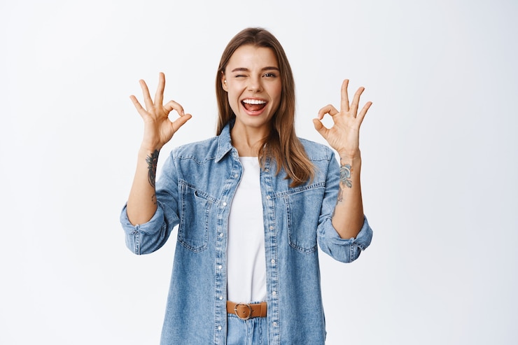 Foto grátis menina sorridente e animada mostrando sinais de ok e piscando, diga sim ou tudo bem, elogie, ótimo trabalho, gesto de bom trabalho, feliz em pé contra uma parede branca