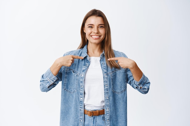 Menina sorridente e confiante apontando para si mesma para se autopromover, valorizar suas próprias realizações, gabar-se de realizações, ficar em pé de branco