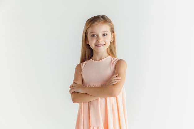 Menina sorridente elegante posando com vestido isolado no fundo branco do estúdio. Modelo feminino loiro branco. Emoções humanas, expressão facial, infância. De pé com as mãos cruzadas.