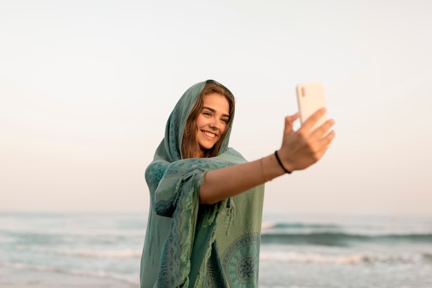 Menina sorridente, embrulhado, em, echarpe, levando, selfie, de, telefone móvel, em, praia