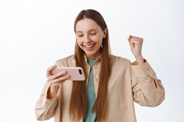 Menina sorridente olha para o smartphone e se alegra, empurra o punho satisfeito, ganhando no videogame para celular, comemorando a vitória online, atingir a meta no aplicativo, em pé sobre a parede branca