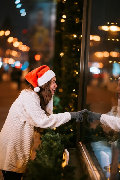 Menina sorridente olhando pela vitrine, as luzes da cidade no fundo, em uma rua de uma cidade europeia, usando chapéu de Papai Noel.