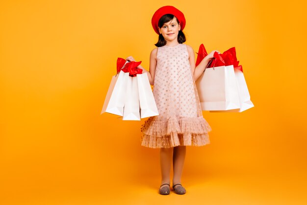 Menina sorridente segurando sacolas de compras. Criança alegre em um vestido de pé na parede amarela.