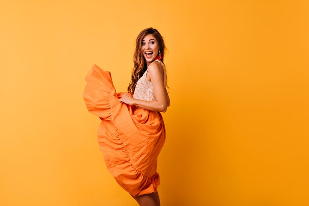 Menina sorridente sensual brincando com sua saia laranja de verão. Foto interna do feliz modelo feminino desfrutando de retratos.