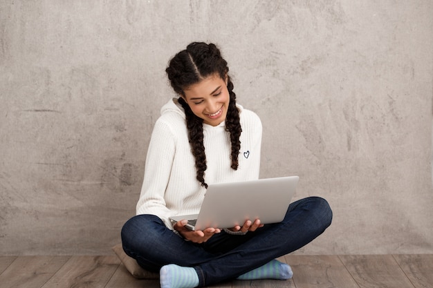 Foto grátis menina sorrindo, segurando laptop, sentada no chão parede bege