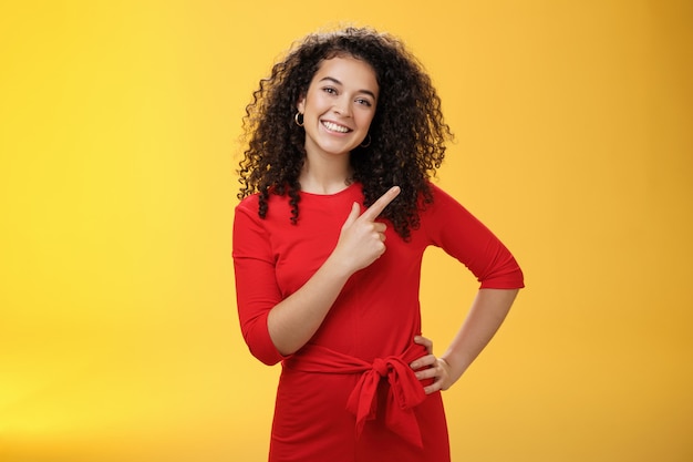 Menina sugerindo experimentar um novo produto. Simpática e agradável jovem morena europeia feliz com cabelos cacheados inclinando a cabeça fofa, sorrindo amplamente, apontando para o canto superior direito para direcionar para o anúncio.