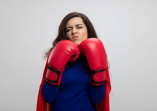 Foto grátis menina super-heroína caucasiana irritada com capa vermelha usando luvas de boxe, parada em um rack isolado na parede branca com espaço de cópia