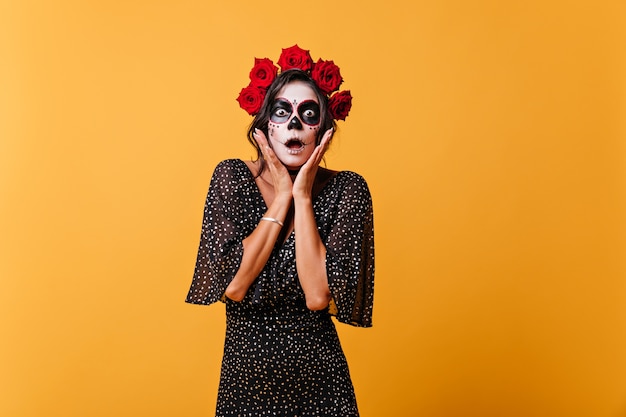 Foto grátis menina surpresa segurou seu rosto e olhou em choque. senhora em roupa preta, coroa rosa e máscara de esqueleto posando na parede laranja.