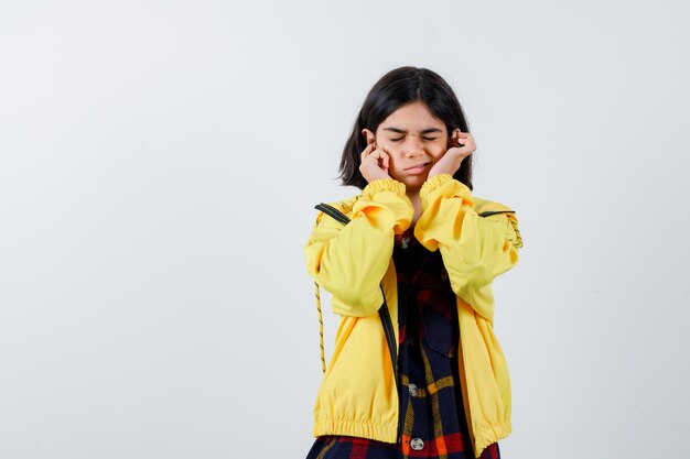 Menina tapando as orelhas com os dedos em camisa quadriculada, jaqueta e parecendo confusa, vista frontal.