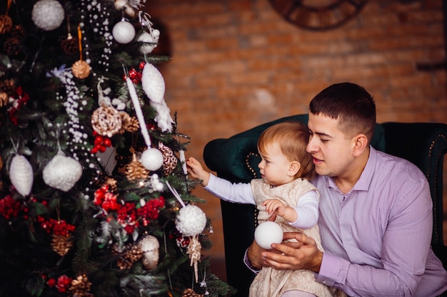 Menina tenta tocar o brinquedo sentado com o pai antes de uma árvore de Natal