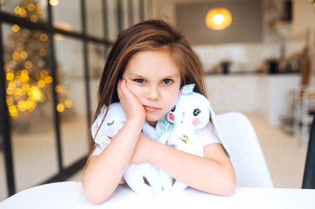 Menina triste abraçando o brinquedo favorito enquanto está sentada à mesa, infância feliz