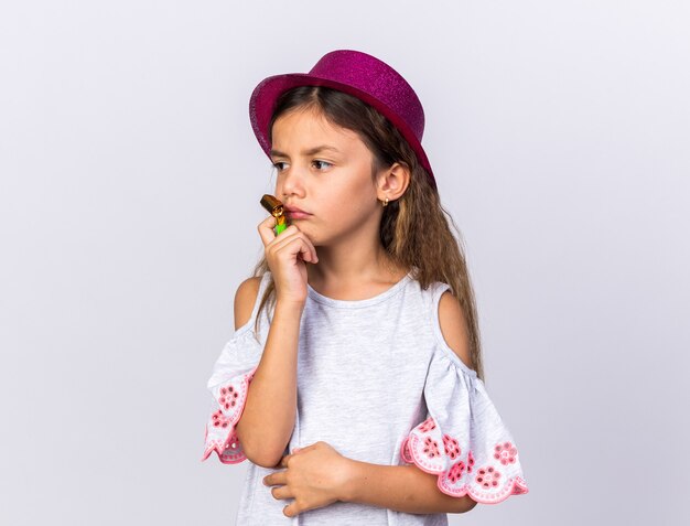 menina triste caucasiana com chapéu de festa roxo segurando o apito de festa e olhando para o lado isolado na parede branca com espaço de cópia