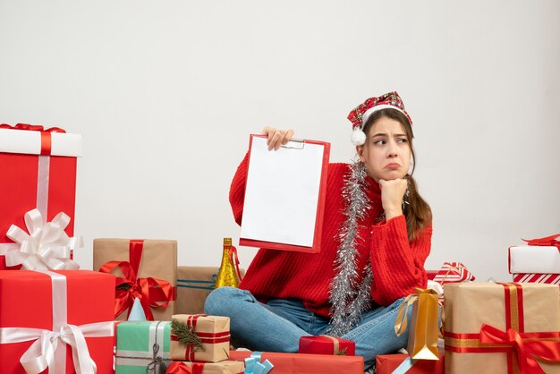 menina triste com chapéu de Papai Noel segurando arquivos sentada em volta de presentes em branco