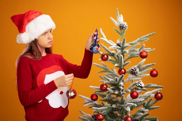 Menina triste com suéter de natal e chapéu de Papai Noel pendurando bolas em uma árvore de Natal e parecendo infeliz sobre um fundo laranja