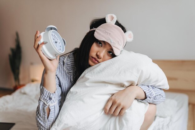 Menina triste de pijama e máscara de dormir está abraçando o travesseiro branco e segurando o despertador