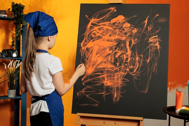 Menina usando aquarela laranja para pintar obras de arte em tela com cavalete, fazendo prática artística para criar design de obra-prima com paleta de aquarela e pincel. criação de pintura com tintura úmida.