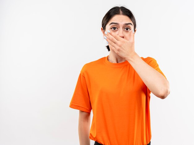 Menina vestindo uma camiseta laranja em choque, cobrindo a boca com a mão em pé sobre uma parede branca