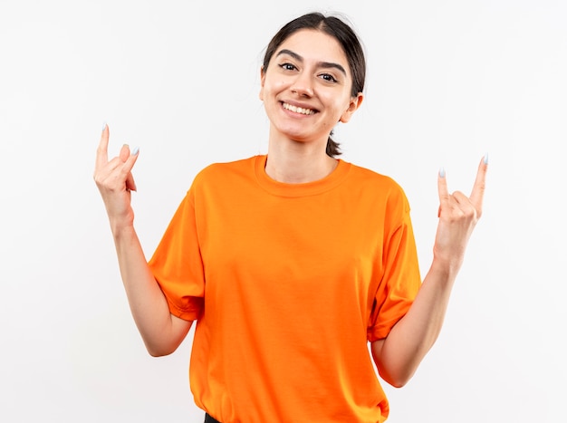 Menina vestindo uma camiseta laranja mostrando o símbolo do rock e sorrindo com uma cara feliz em pé sobre uma parede branca