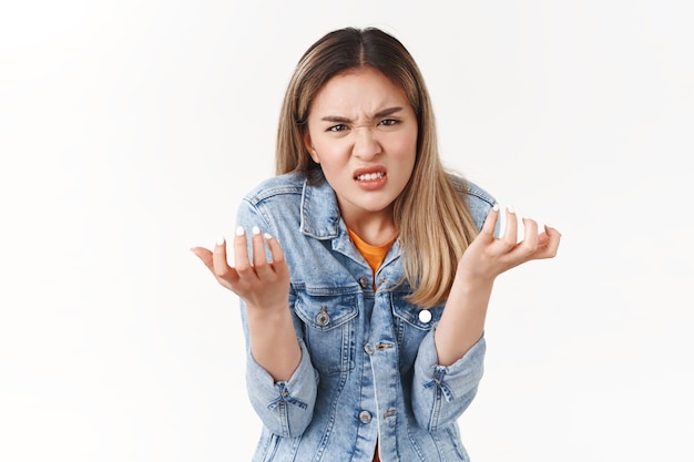 Menina xingando maldição cruel vida injusta perdendo chance participar do evento. Chateado, intenso, irritado, odioso, garota loira asiática, encolhendo-se, fazendo careta, raiva, apertando os punhos irritado, parede branca de pé