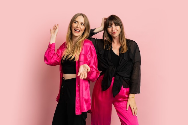 Meninas da moda felizes e sorridentes, vestindo roupas coloridas e elegantes, posando na parede rosa