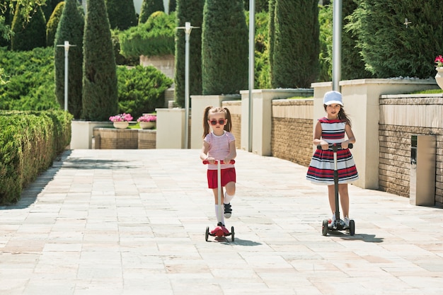 Meninas em dia de sol. Meninas em idade pré-escolar andando de scooter ao ar livre. Crianças felizes e fofas brincando na rua