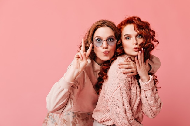 Meninas europeias em êxtase, mostrando o símbolo da paz. foto de estúdio de duas senhoras sonhadoras, abraçando-se no fundo rosa.