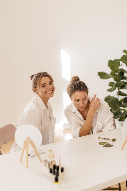 Meninas loiras caucasianas bonitas usam creme para o rosto enquanto estão sentadas no salão de beleza. Conceito de preservar a juventude