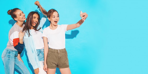 Meninas, tirando fotos de auto-retrato de selfie no smartphone. Modelos posando perto de parede azul no estúdio, fêmea, mostrando emoções de rosto positivo