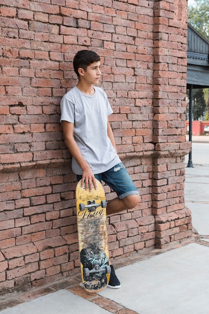 Menino adolescente, inclinar-se, parede tijolo, segurando, skateboarding, olhando
