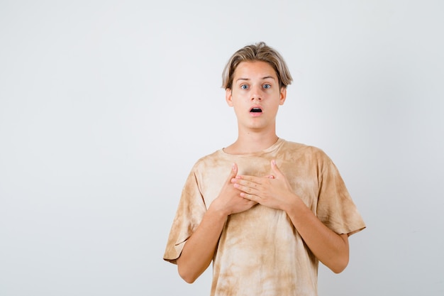 Menino adolescente mantendo as mãos no peito em t-shirt e parecendo perplexo, vista frontal.