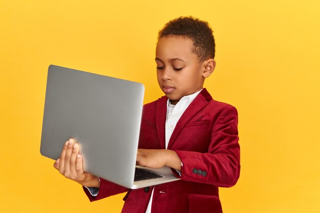 Menino africano bonito e elegante digitando em um dispositivo eletrônico portátil genérico, enviando mensagens online através de redes sociais, aprendendo remotamente. Infância, tecnologia, comunicação e educação
