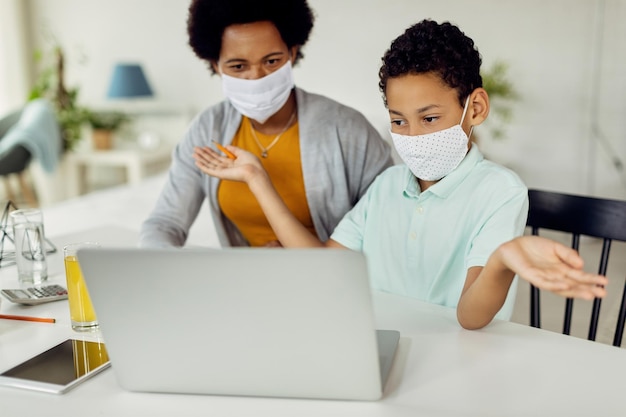 Menino afro-americano confuso usando laptop enquanto estudava em casa com sua mãe durante o bloqueio do cvovid-19.