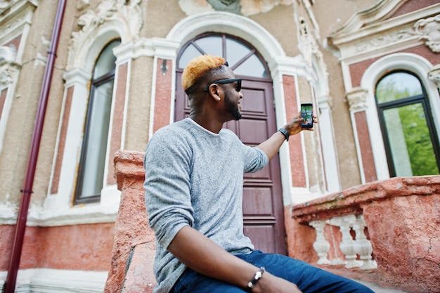 Menino afro-americano elegante na camisola cinza e óculos de sol pretos posou na rua Cara negro na moda fazendo selfie no telefone