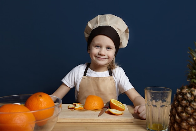 Menino alegre usando avental e chapéu de chef em pé na cozinha moderna, cozinhando salada de frutas. retrato de uma criança do sexo masculino, branca e fofa, de uniforme, fazendo suco fresco, cortando e descascando laranjas