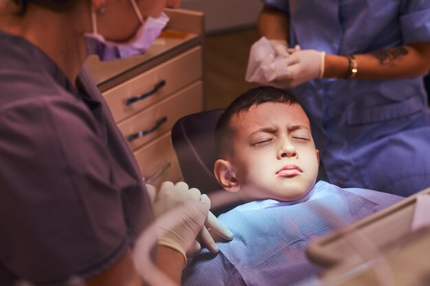 Menino assustado está sentado na cadeira de dentista, esperando o início do tratamento. Há dentista e seu assistente.