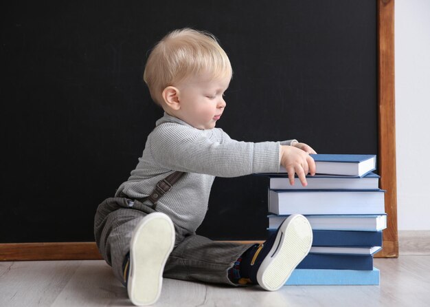 Menino bonitinho sentado no chão perto do quadro-negro com livros