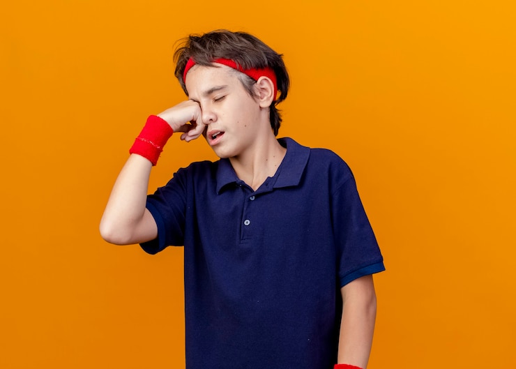 Foto grátis menino bonito e esportivo dolorido usando bandana e pulseiras com aparelho dentário, colocando a mão no olho com os olhos fechados, isolado na parede laranja com espaço de cópia