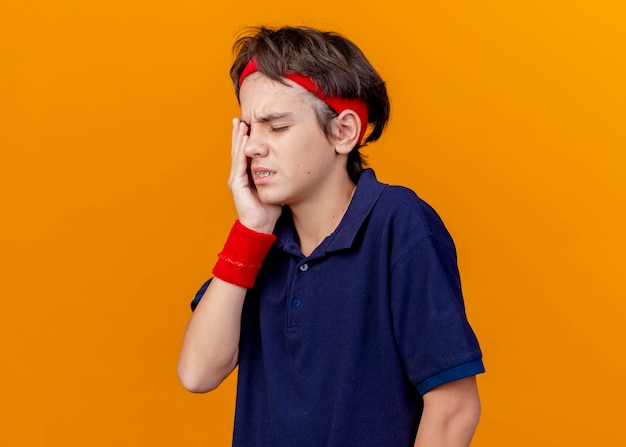 Menino bonito e esportivo dolorido usando bandana e pulseiras com aparelho dentário, mantendo a mão no olho isolado na parede laranja