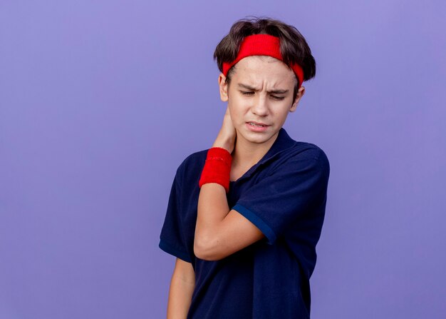 Menino bonito e esportivo dolorido usando bandana e pulseiras com aparelho dentário olhando para baixo, mantendo a mão atrás do pescoço isolada na parede roxa