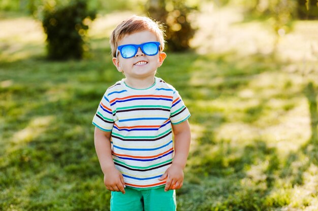 Menino bonito sorri no jardim de verão em óculos de sol brilhantes e mostra sua felicidade.