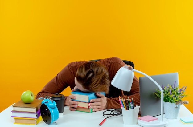 Menino cansado de jovem estudante sentado à mesa com ferramentas escolares colocando a cabeça em livros isolados na parede amarela