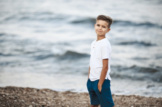 Menino caucasiano está de pé na praia, perto do mar ondulado, vestido com camiseta branca e calção azul