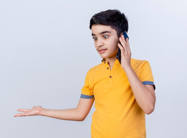 Menino caucasiano impressionado olhando para baixo, falando no telefone, mostrando a mão vazia isolada no fundo branco